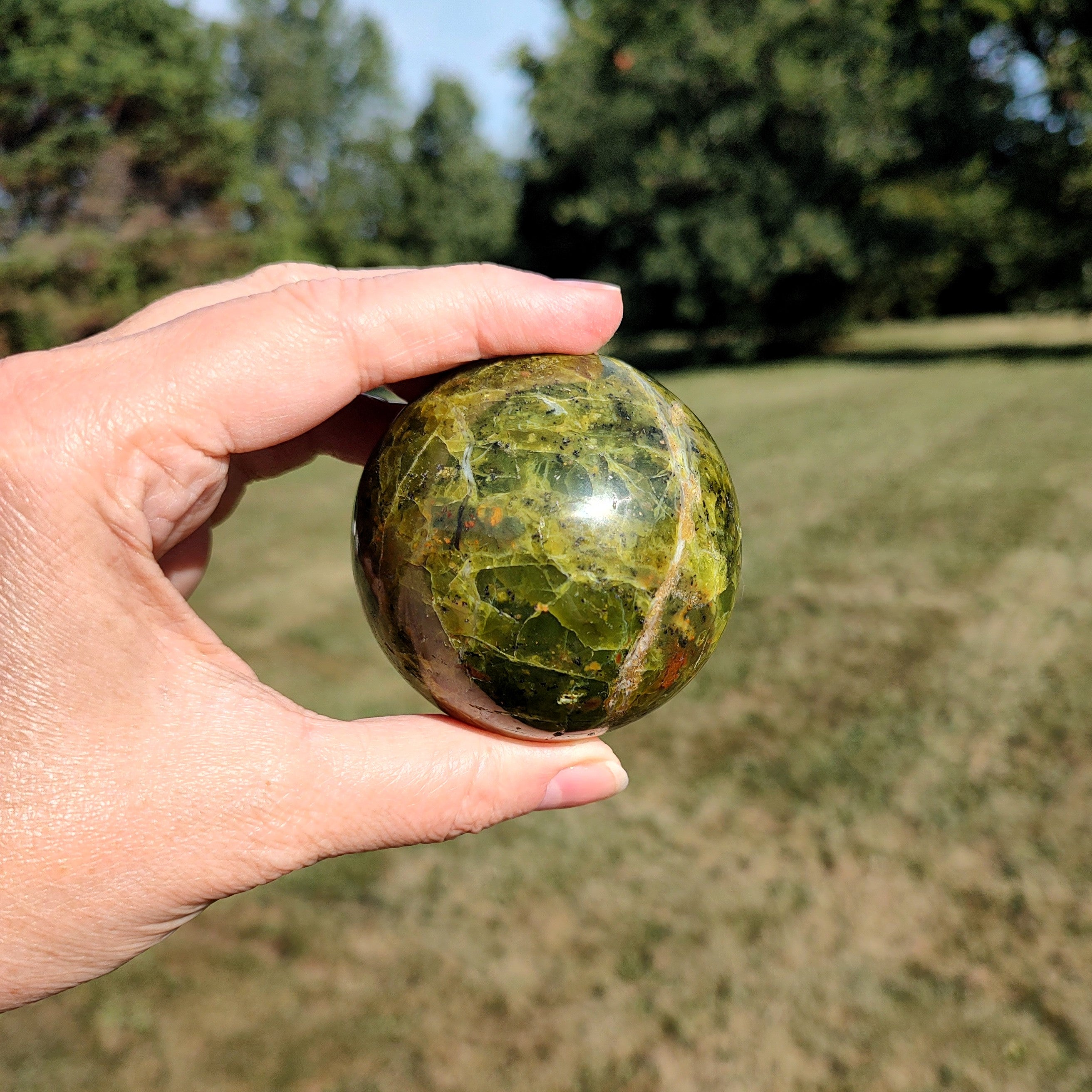 Hand holding a green opal sphere outdoors with trees in the background.