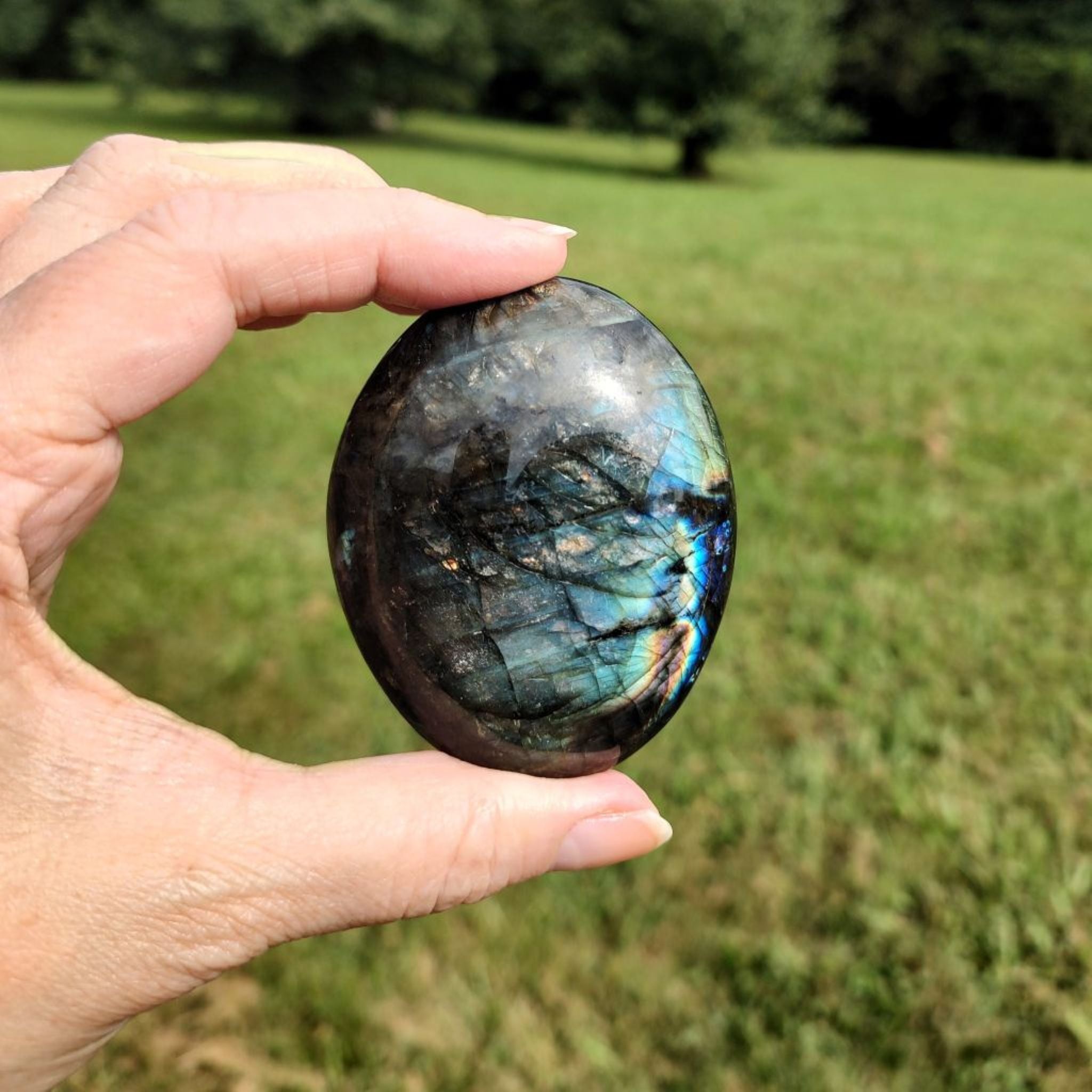 Hand holding a labradorite palm stone in direct sunlightm with a blurred green field background.