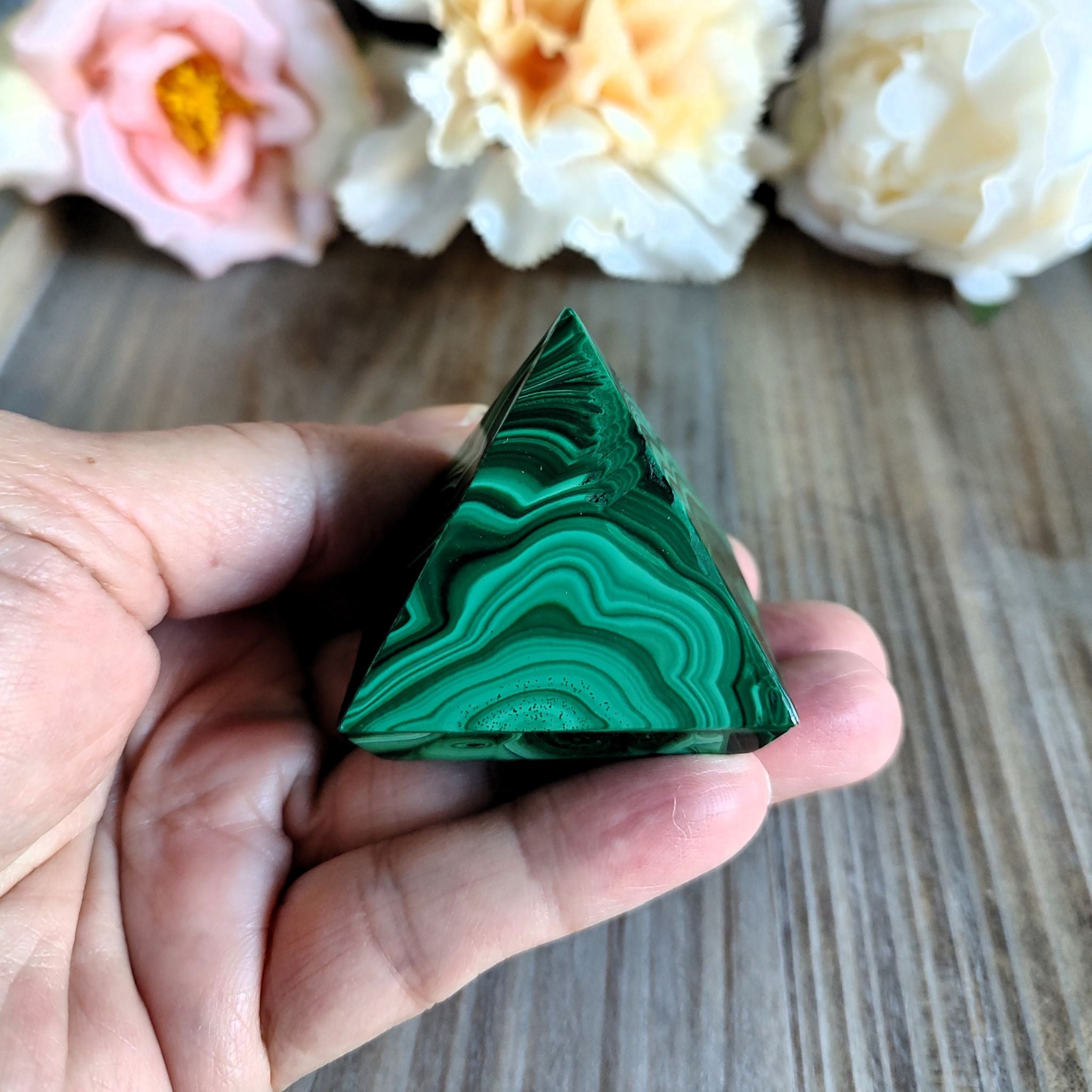 Hand holding a handcrafted malachite pyramid  in front of a wooden surface with flowers in the background. 