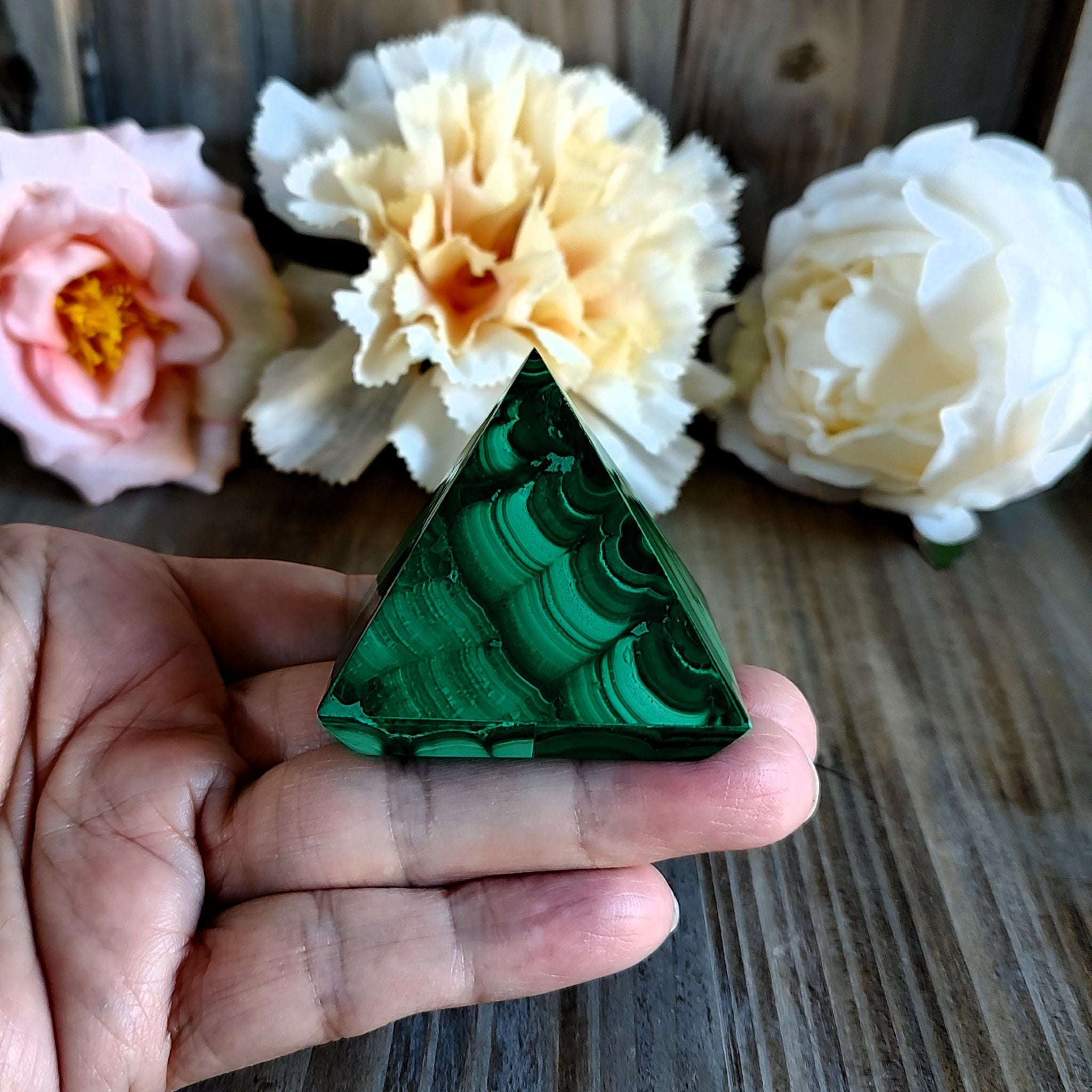 Hand holding a handcrafted malachite pyramid  in front of a wooden surface with flowers in the background. 