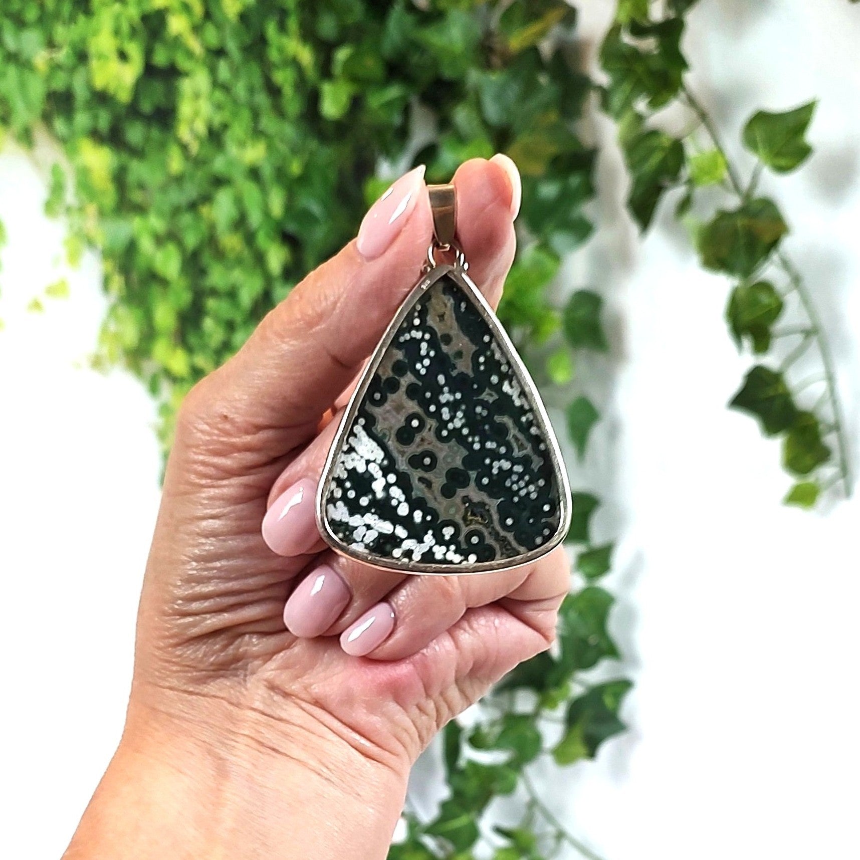 Hand holding a teardrop-shaped ocean jasper pendant against a leafy green background. 