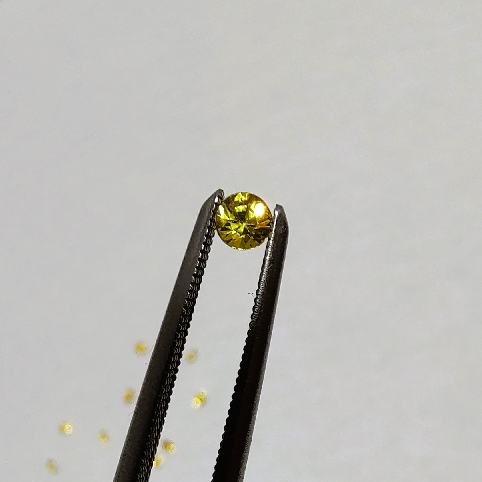 Round faceted yellow sapphire held by tweezers on a light gray background.